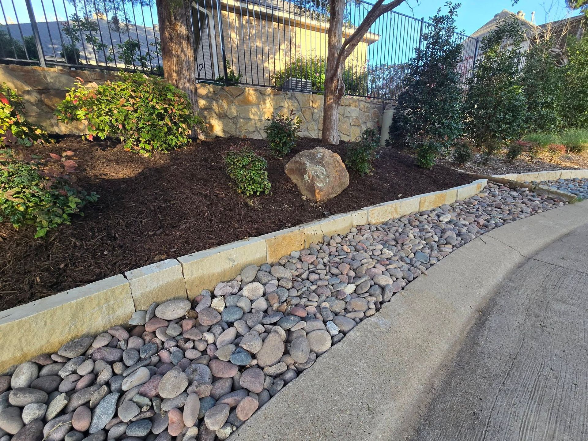 Landscaped roadside bed with rocks, mulch, and a curved concrete curb beside a fence