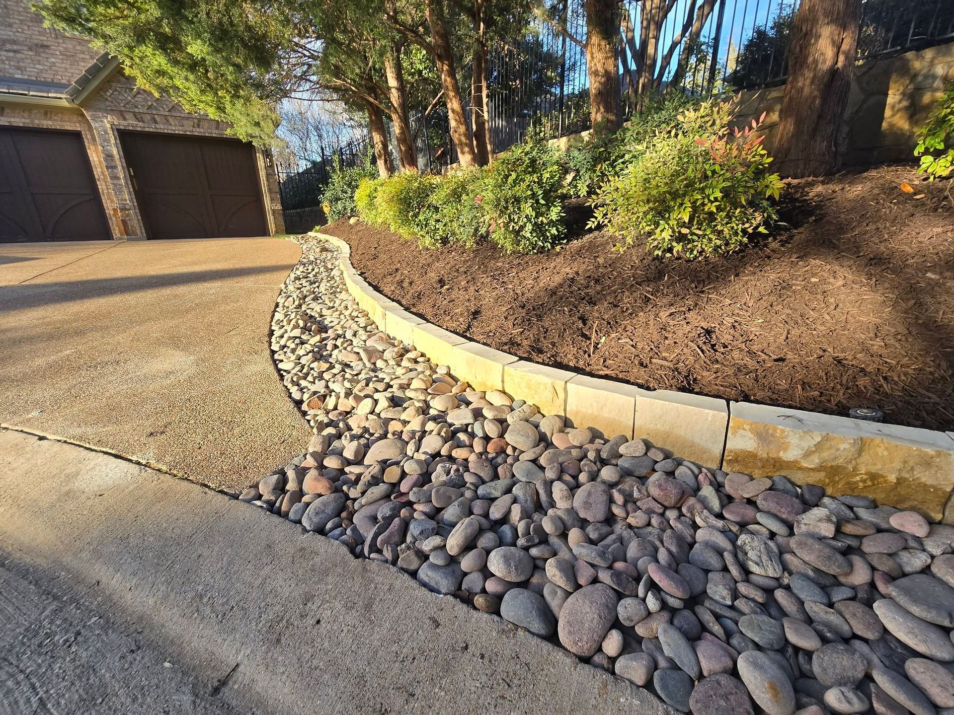 Curved rock-lined garden bed beside a concrete driveway with shrubs and trees