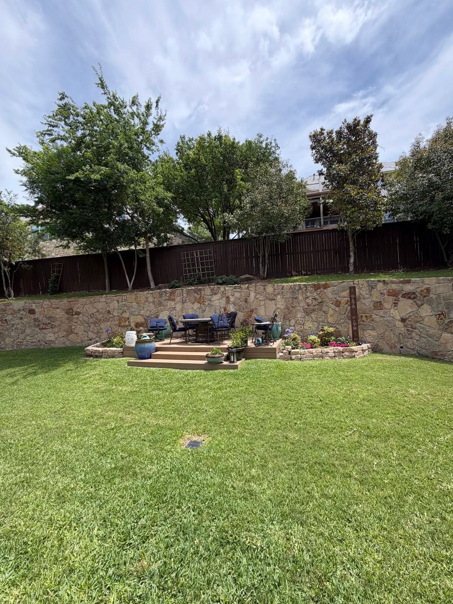 Backyard lawn with a raised stone patio, patio furniture, flower beds, and a wooden fence under a cloudy sky