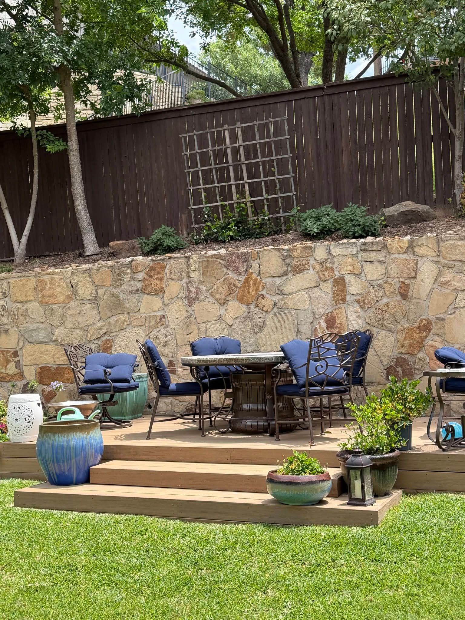 Backyard patio with blue chairs, a table, potted plants, stone wall, and green lawn