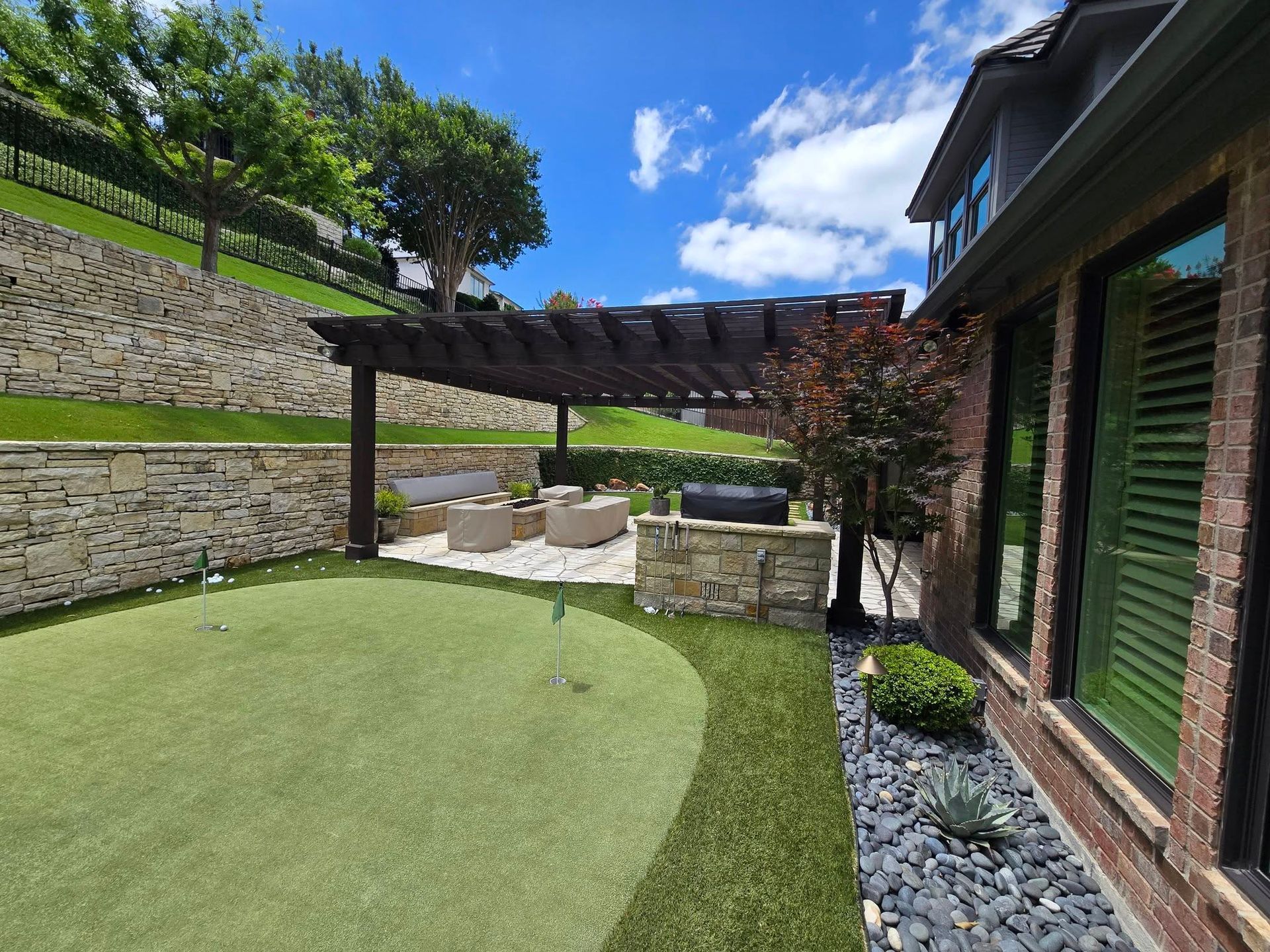 Backyard patio with pergola, stone wall, and artificial lawn beside a brick house