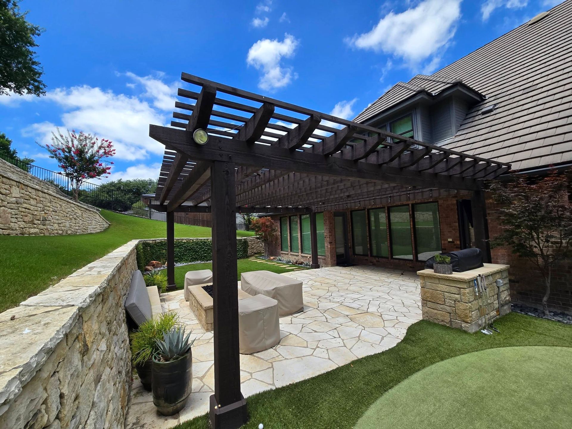 Stone patio with pergola beside a brick house, landscaped lawn, and blue sky