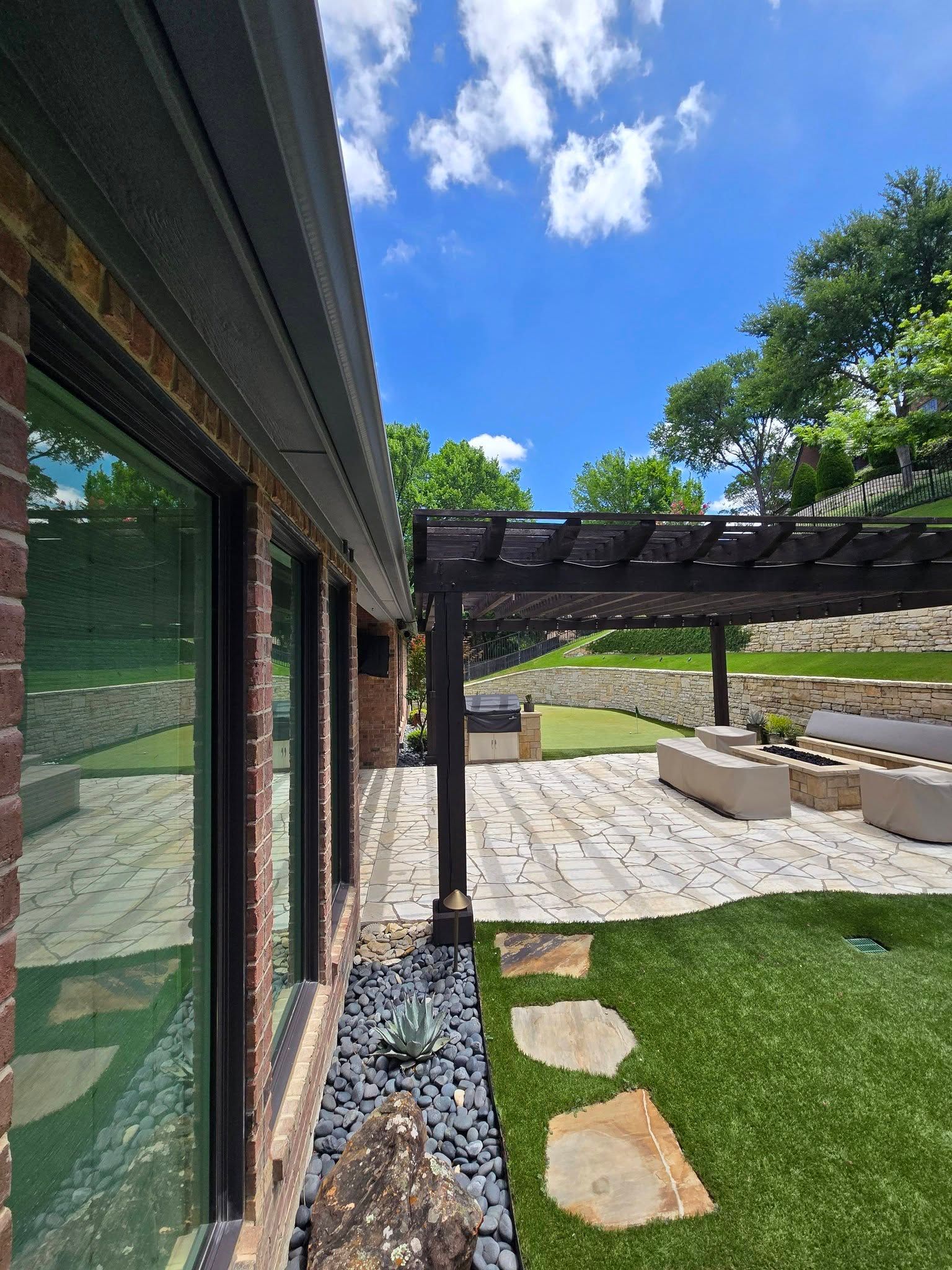 Modern backyard patio with pergola, stepping stones, and green lawn beside a brick house