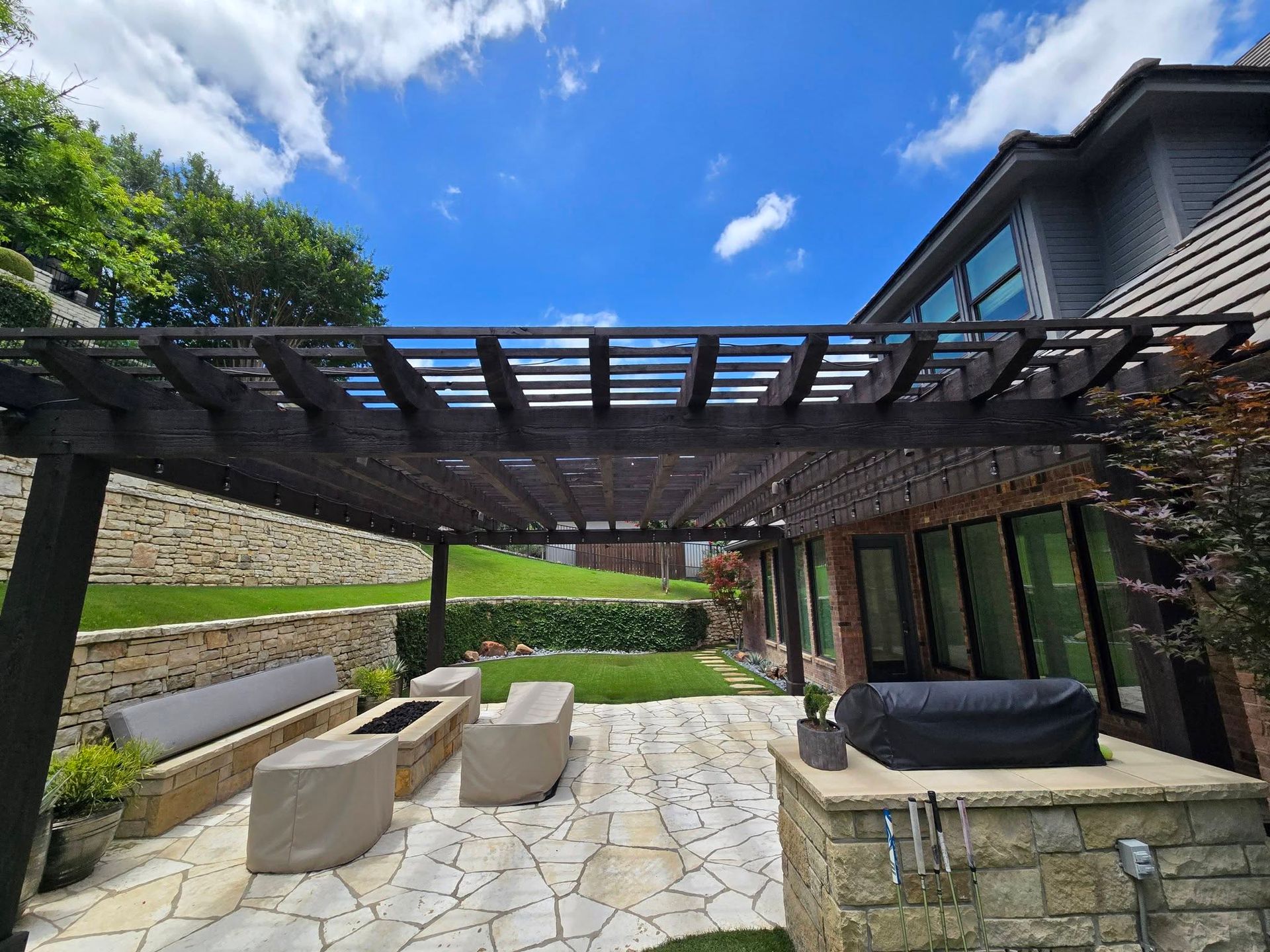 Stone patio with pergola, outdoor seating, and glass doors beside a modern house under a blue sky