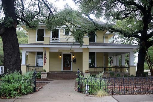 Yellow two-story house with porch and trees, path to the front door, and a black fence.