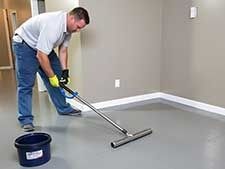 Man applying epoxy floor coating with a roller, bucket beside him, in a room.