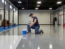 Man kneeling, cleaning shiny gray floor in gym; blue bucket beside him.