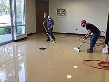 Two people cleaning a shiny, tan floor with industrial vacuum equipment. Large window and door in the background.