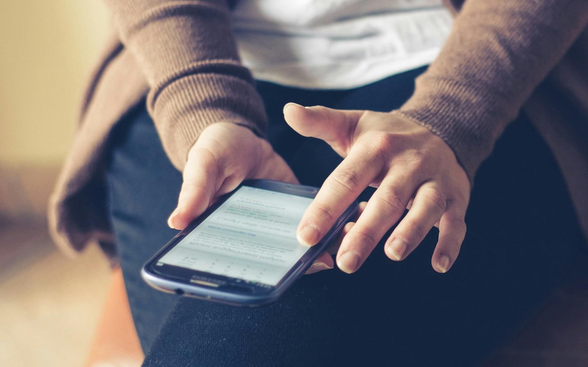 Person holding a smartphone, tapping the screen with a finger. Wearing a brown sweater and blue jeans.