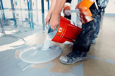 Person pouring gray liquid from a red bucket onto a floor during a flooring project.