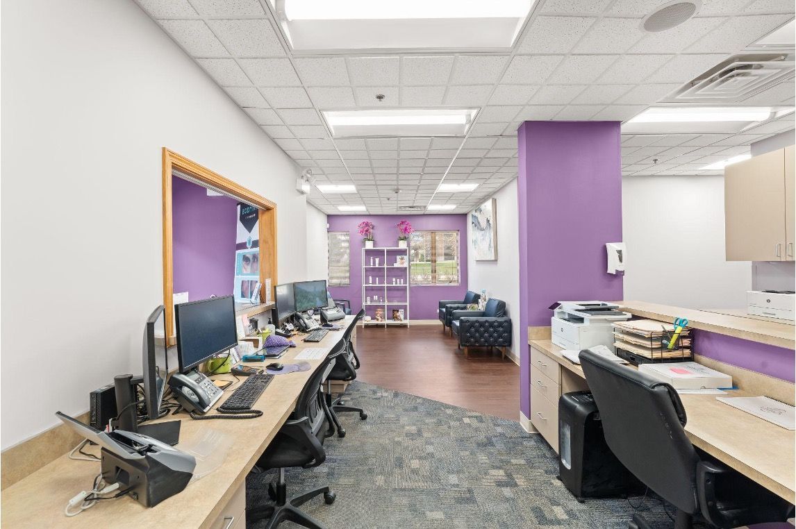 Bright office reception area with desks, computer monitors, purple accent walls, and carpeted floor.