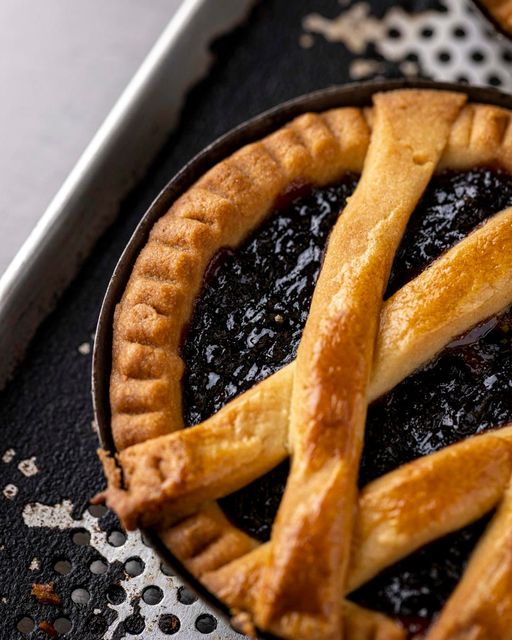Primo piano di una crostata ripiena di marmellata scura, ricoperta da una grata dorata e posta su una teglia nera.