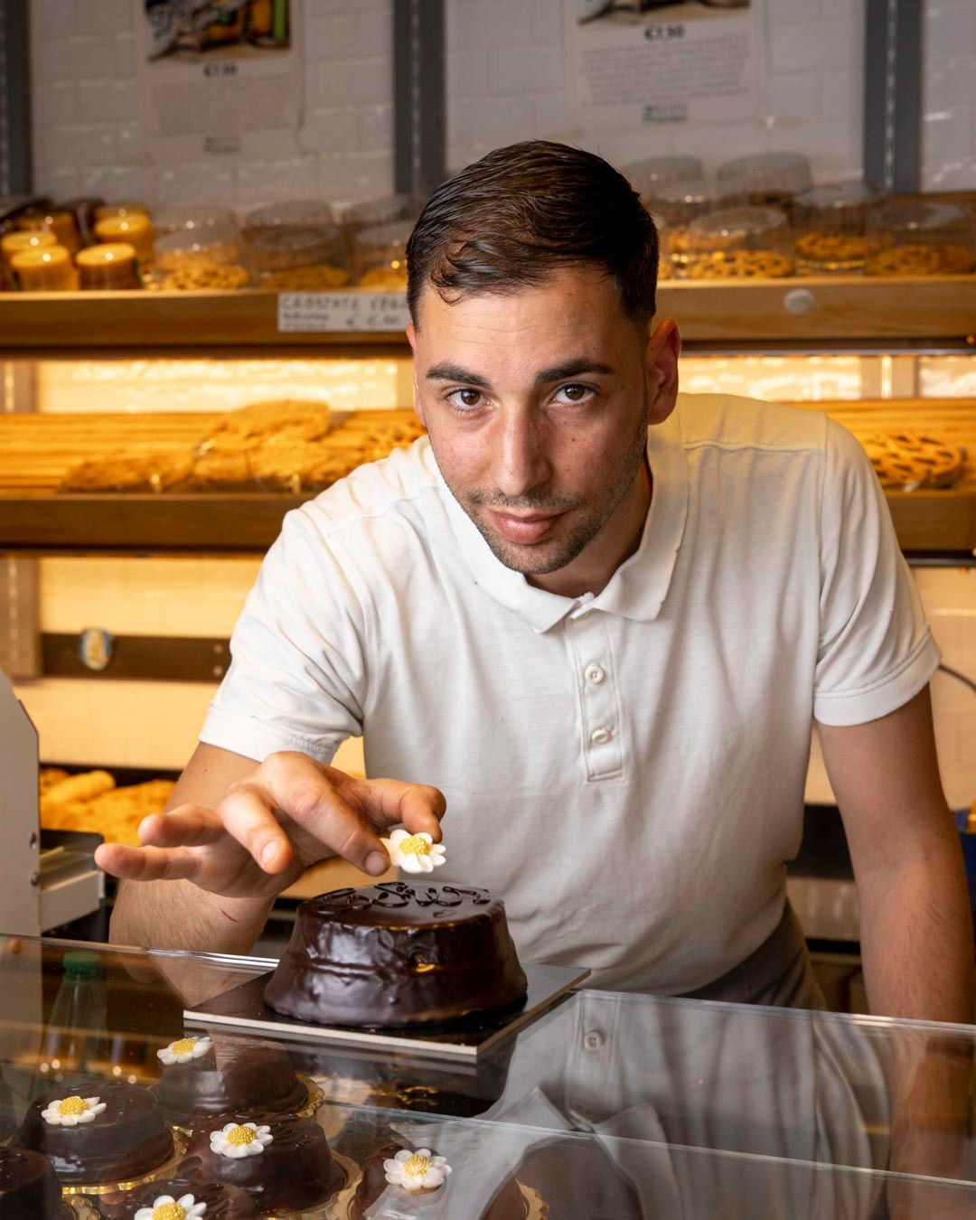 Un fornaio posiziona un fiore decorativo su una torta al cioccolato dietro un bancone di vetro in una panetteria.