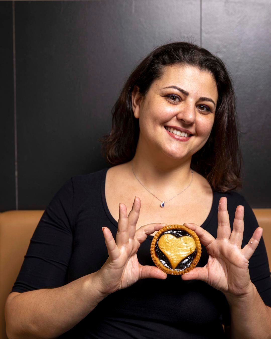 Donna sorridente che tiene in mano un biscotto a forma di cuore. Top nero, sfondo marrone.