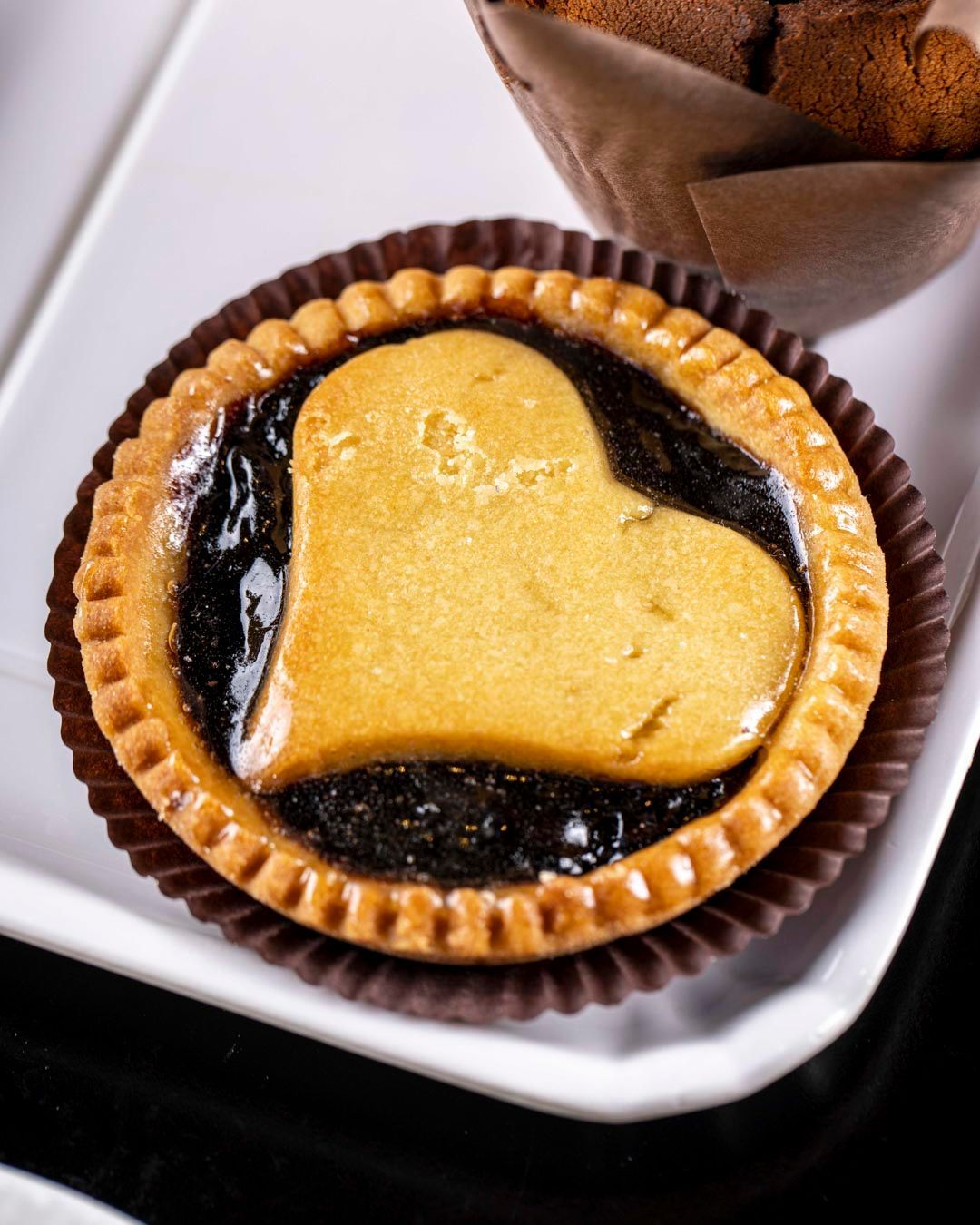 Crostata di pasta sfoglia a forma di cuore con ripieno scuro, adagiata su un piatto bianco, con un muffin sullo sfondo.