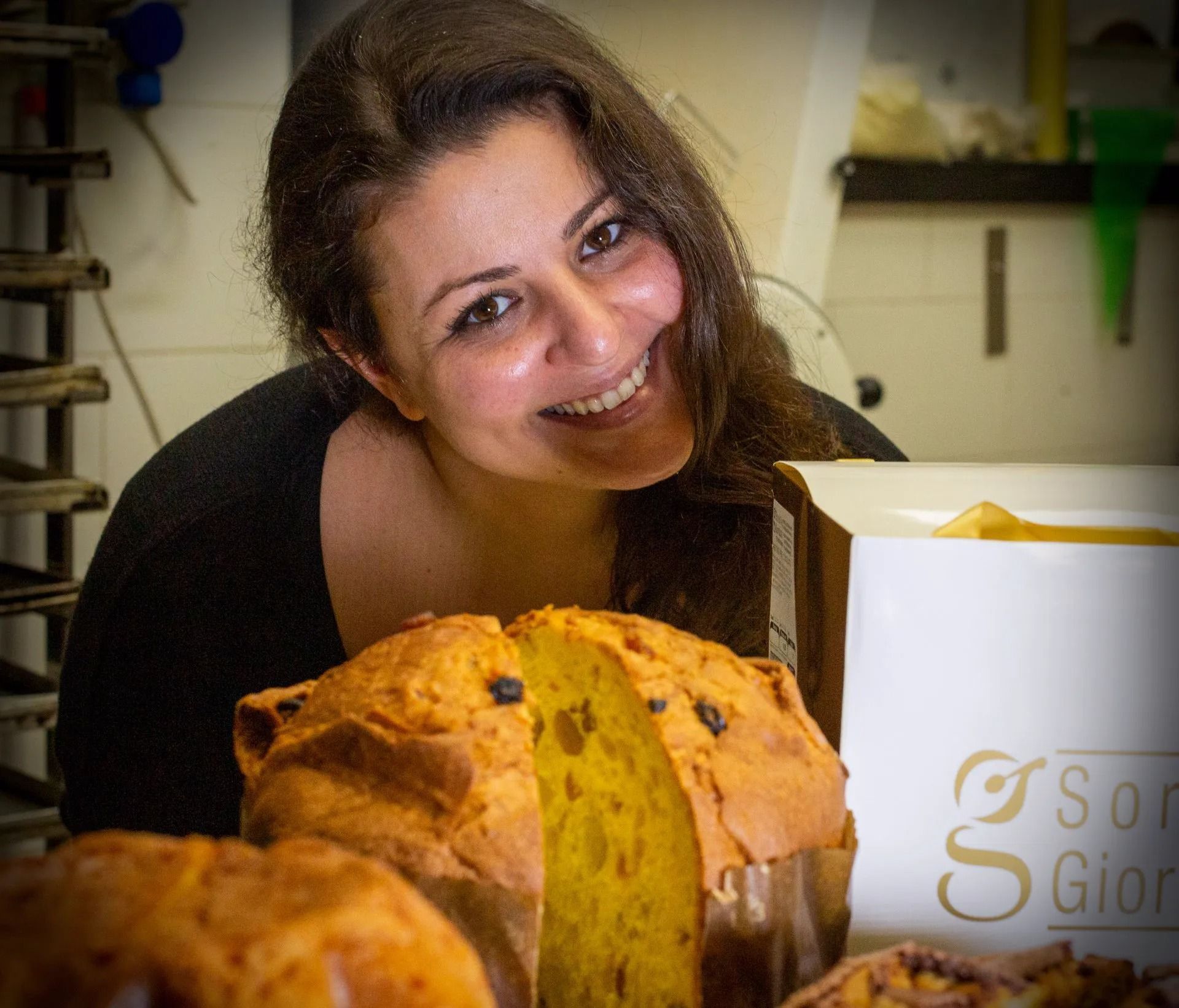 Donna sorridente dietro un panettone parzialmente tagliato con il logo, in un ambiente da panetteria.