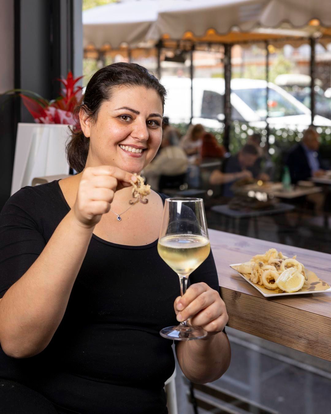 Una donna sorride, tenendo in mano dei calamari fritti e un bicchiere di vino bianco in un ristorante.
