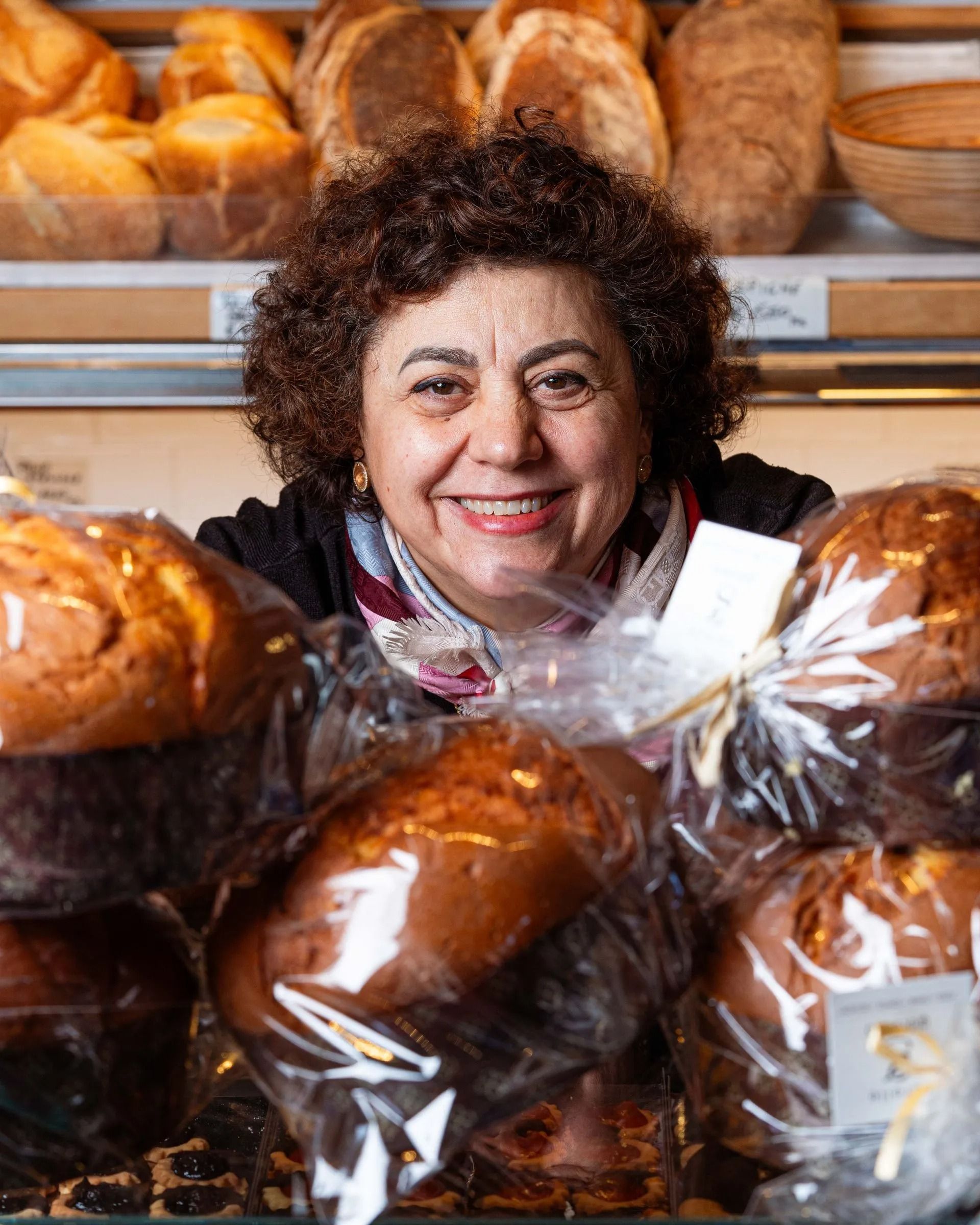 Donna sorridente, dietro prodotti da forno confezionati in un panificio, con pane sullo sfondo.