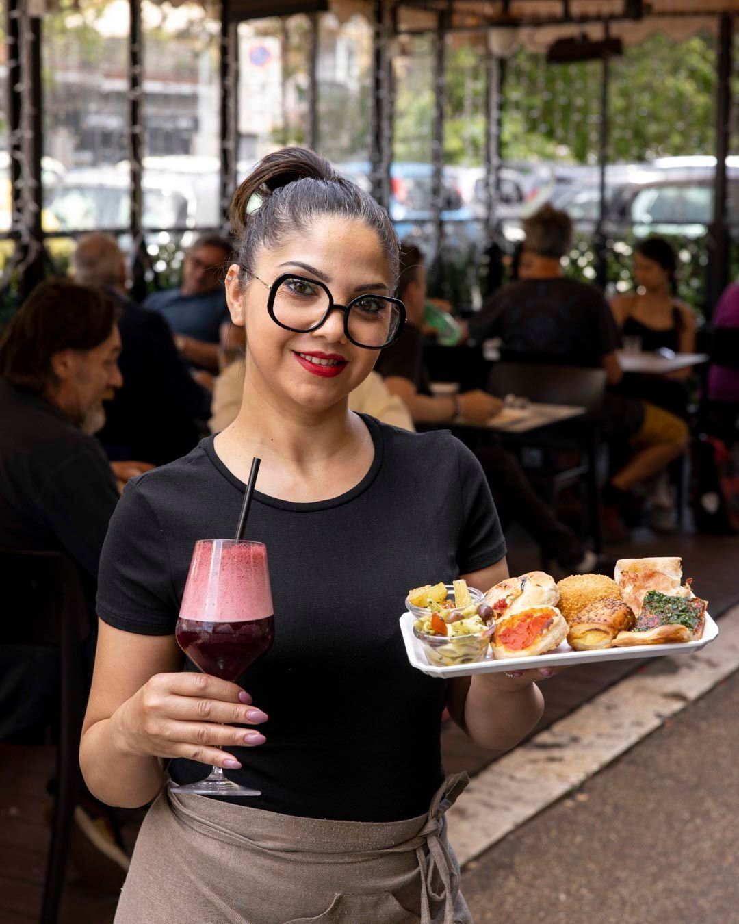 Una donna con il grembiule tiene in mano un vassoio con del cibo e una bevanda in un bar all'aperto.