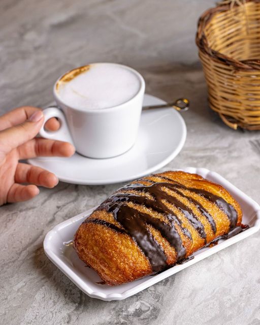 Persona che prende una tazza di caffè, accanto a un dolce ricoperto di cioccolato su un piatto bianco.