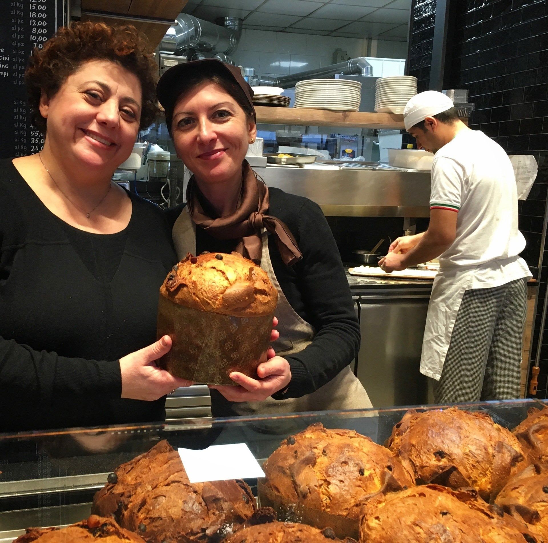 Due donne in un panificio tengono in mano una pagnotta appena sfornata. Un altro fornaio lavora dietro il bancone; esposizione di prodotti da forno.