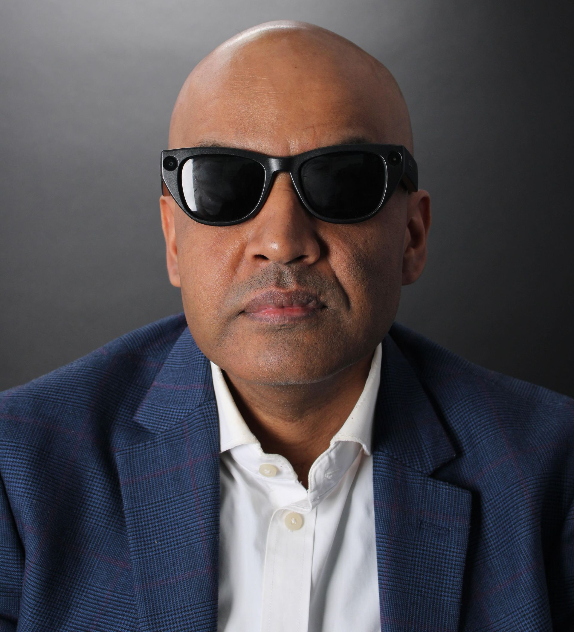 A head and shoulders picture of a man wearing a navy blue jacket with a white shirt & black sun glasses.