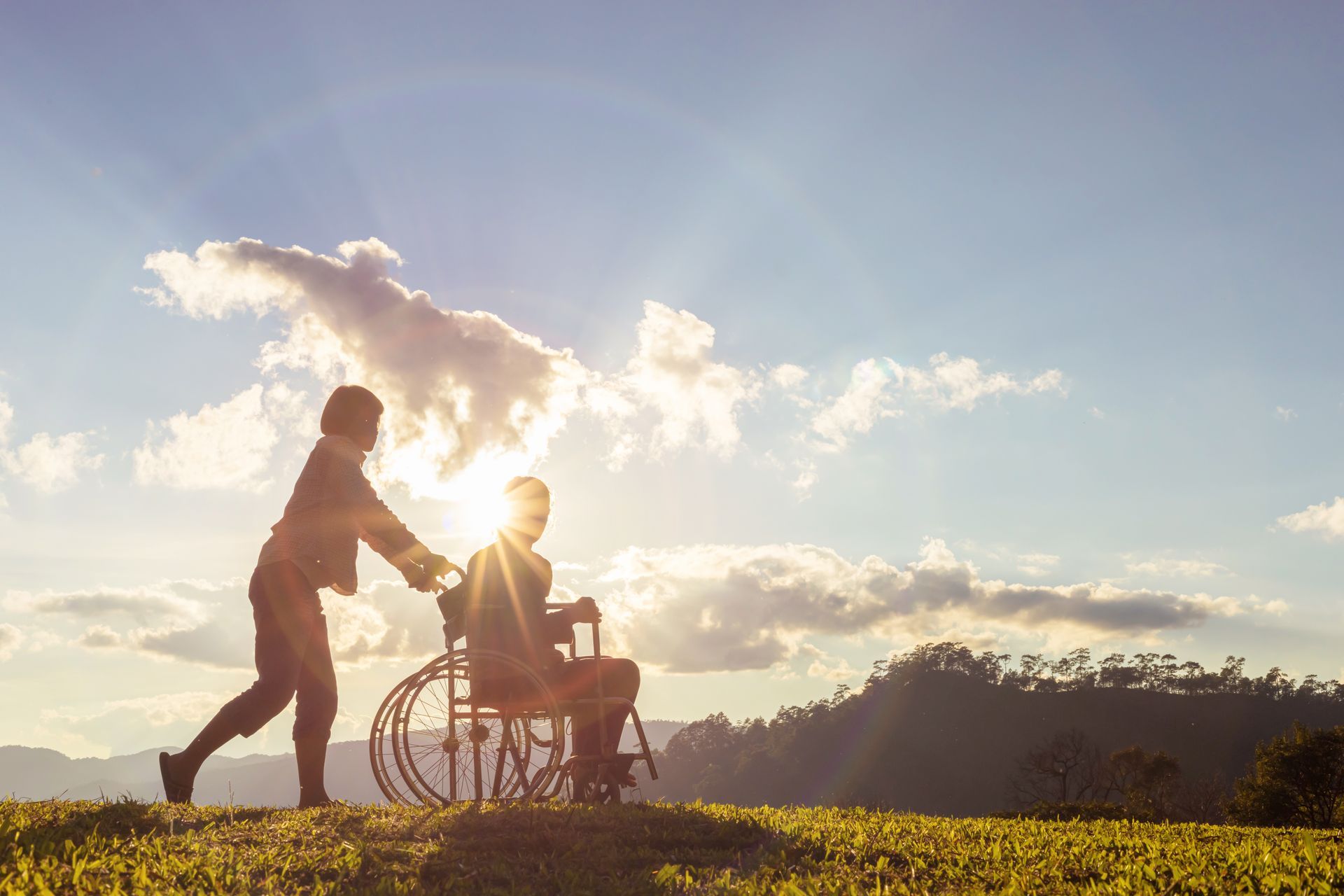 A woman pushing a man in a wheel chair out side in the country side