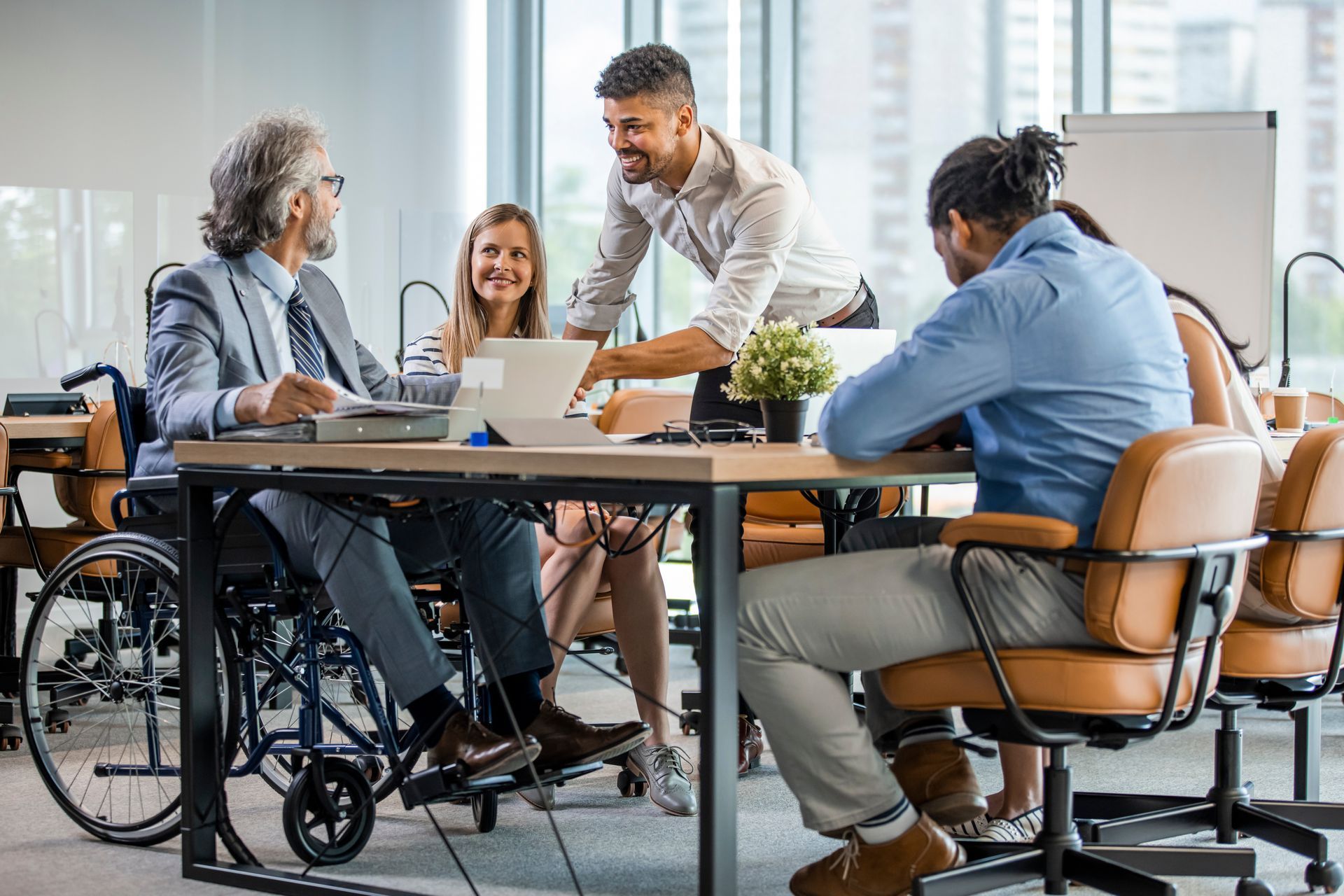 Office environment - Inclusive disability within the work place. Men & woman sat around a office desk with one man in a wheel chair working.