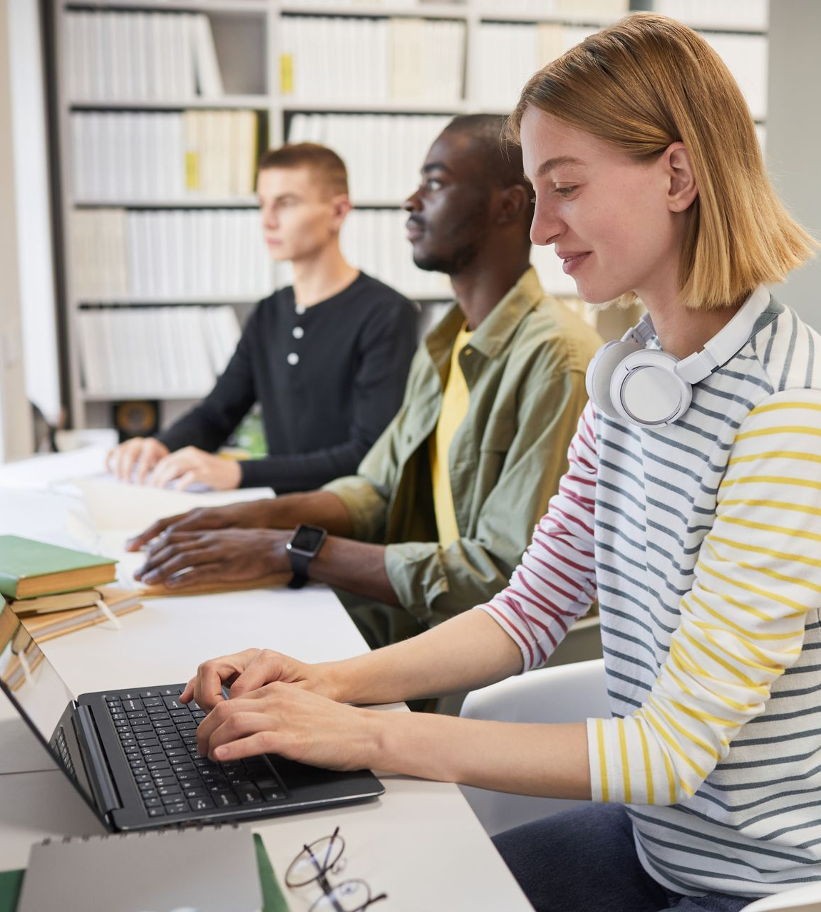 Two men reading braille with their hands at a desk, whilst a lady beside them is using a braille lap top with ear phone around her neck.