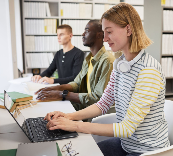 Two men reading braille with their hands at a desk, whilst a lady beside them is using a braille lap top with ear phone around her neck.