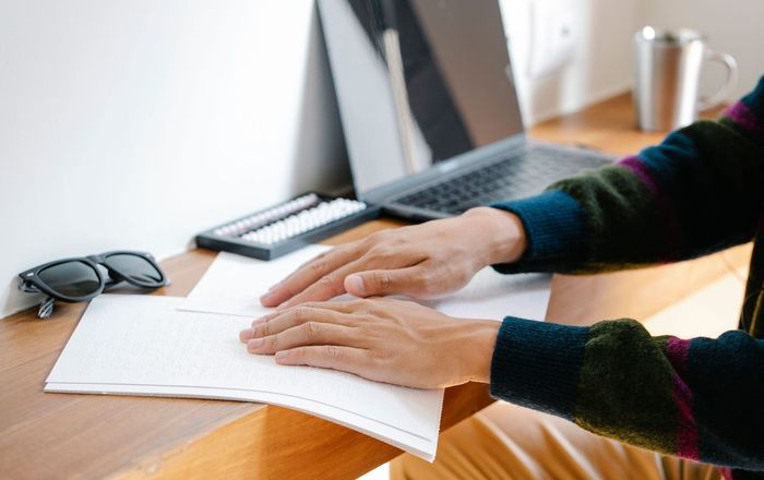 Man sat at a thin wooden long bench table reading braille. On the work bench is a black pair of glasses, braille keyboard, lap top & silver coffee mug.