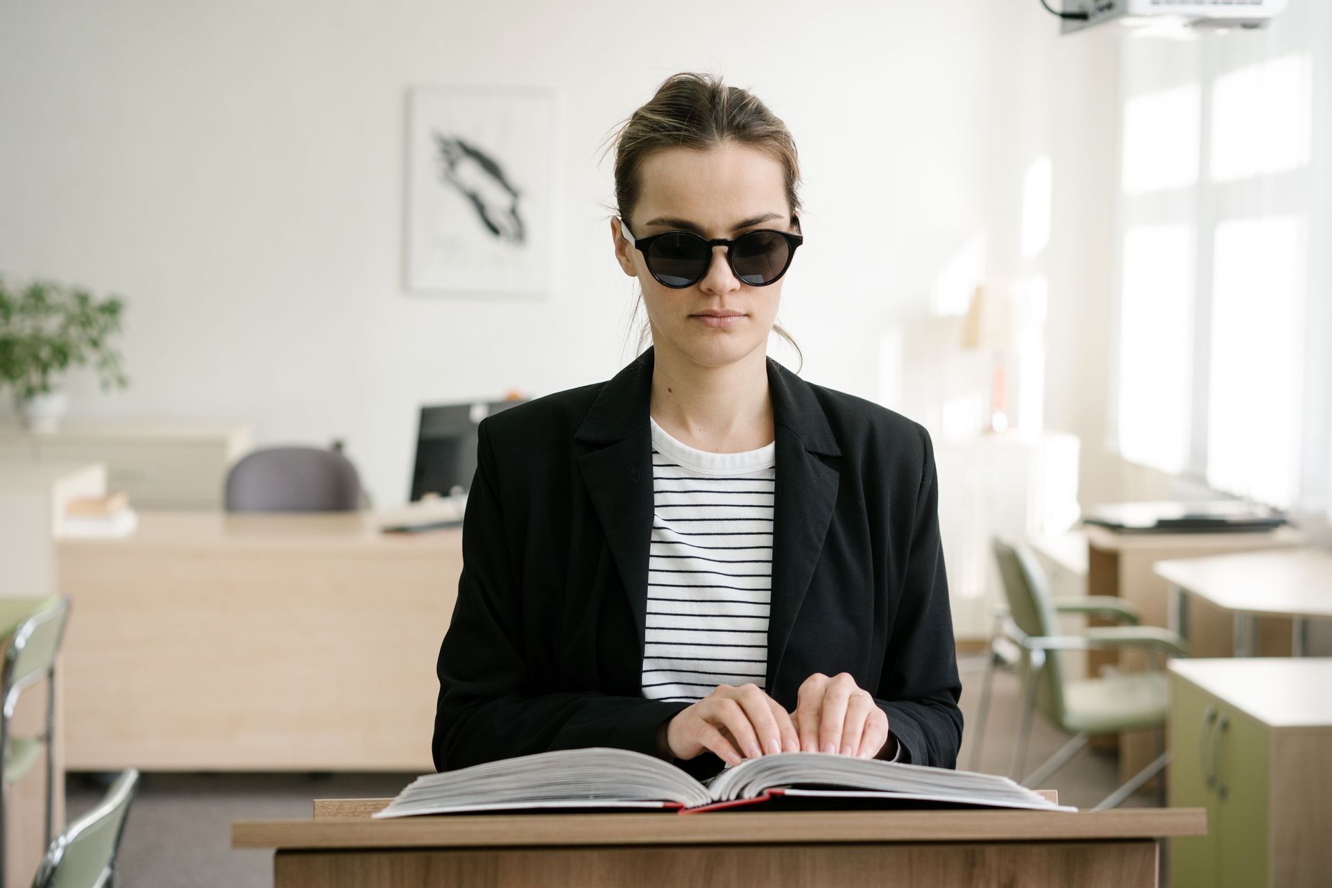 Lady sat in a office at a work desk reading braille with a black suit jacket, white & black stripe top, dark glasses & her hair tied back.