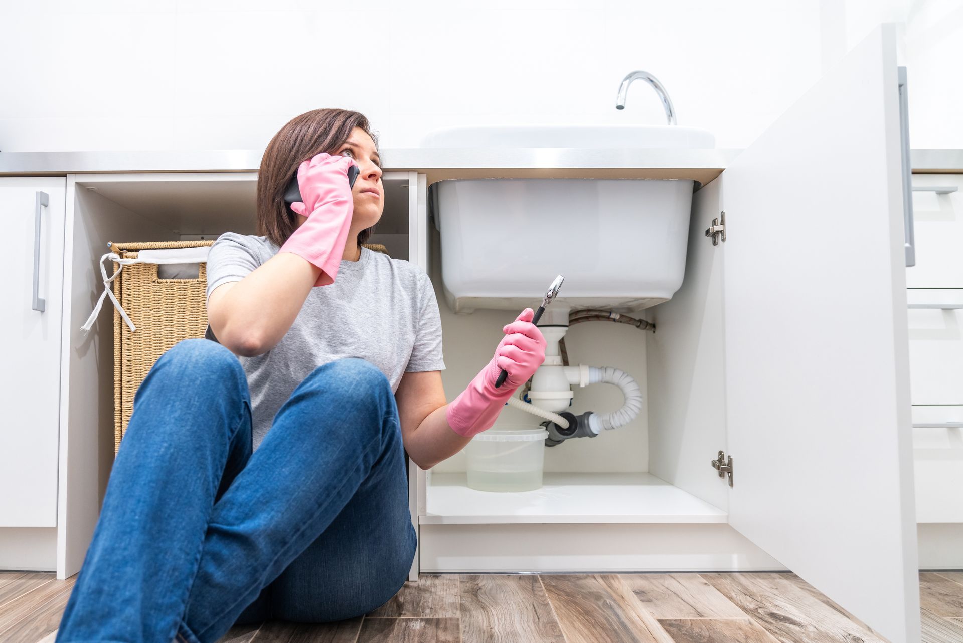 A woman on the phone by a leaking sink is calling for emergency plumbing services. A woman on the phone by a leaking sink is calling for emergency plumbing services.
