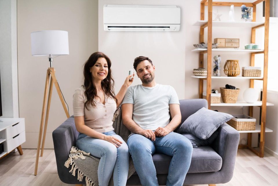 Couple sitting on a gray sofa in a bright living room with a wall-mounted air conditioner and wooden shelves