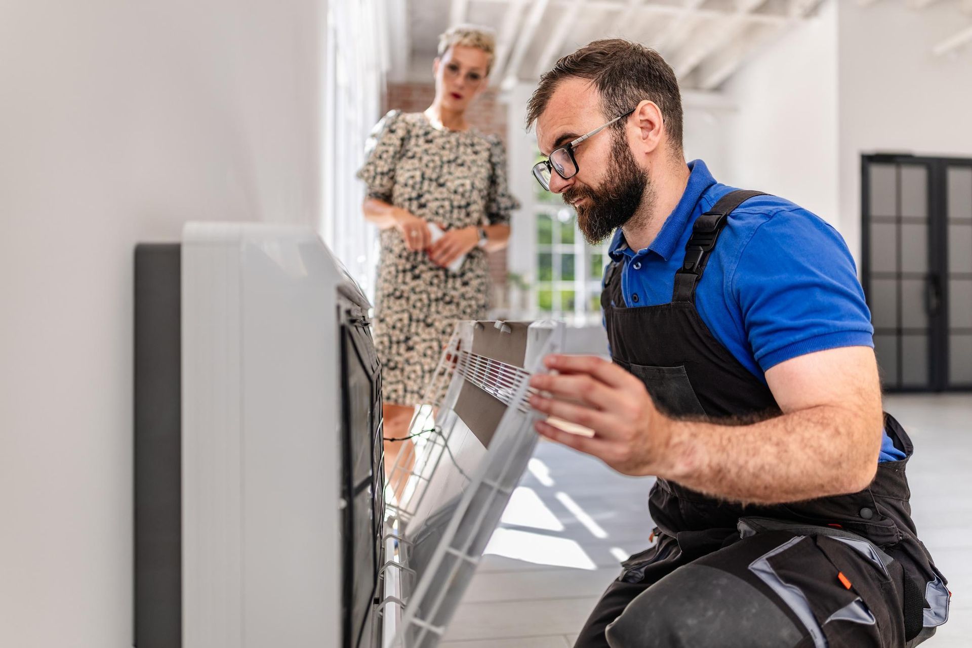 Technician kneeling beside a wall-mounted unit while a woman watches in a bright office hallway