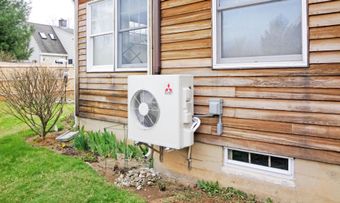Outdoor heat pump unit mounted on the side of a wooden house beneath a window