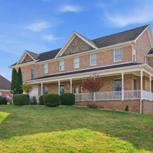 Brick suburban house with white porch railing, manicured shrubs, and a sloped green lawn under blue sky
