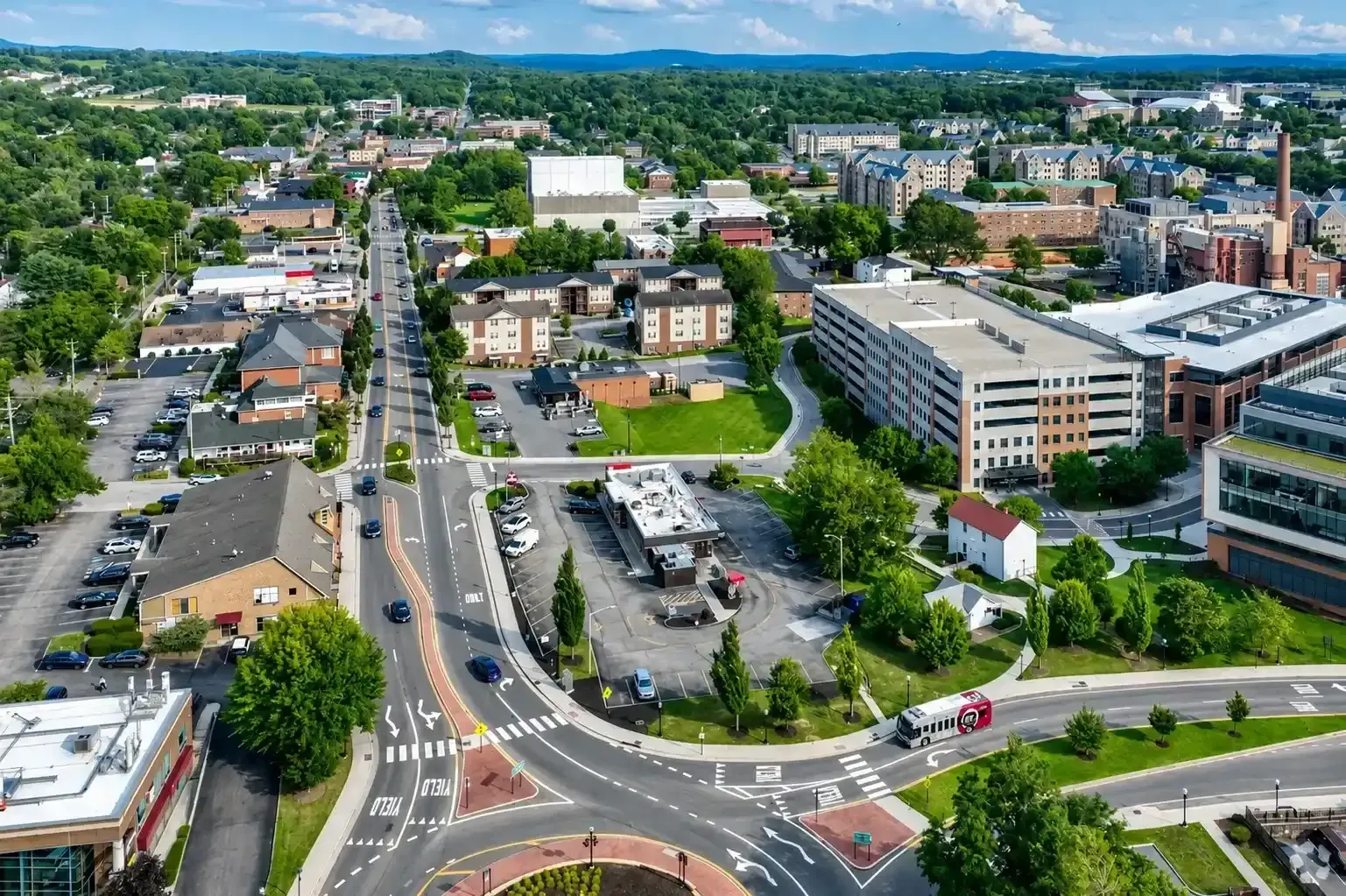 Aerial view of a city intersection with cars, buildings, and tree-lined streets under a clear sky