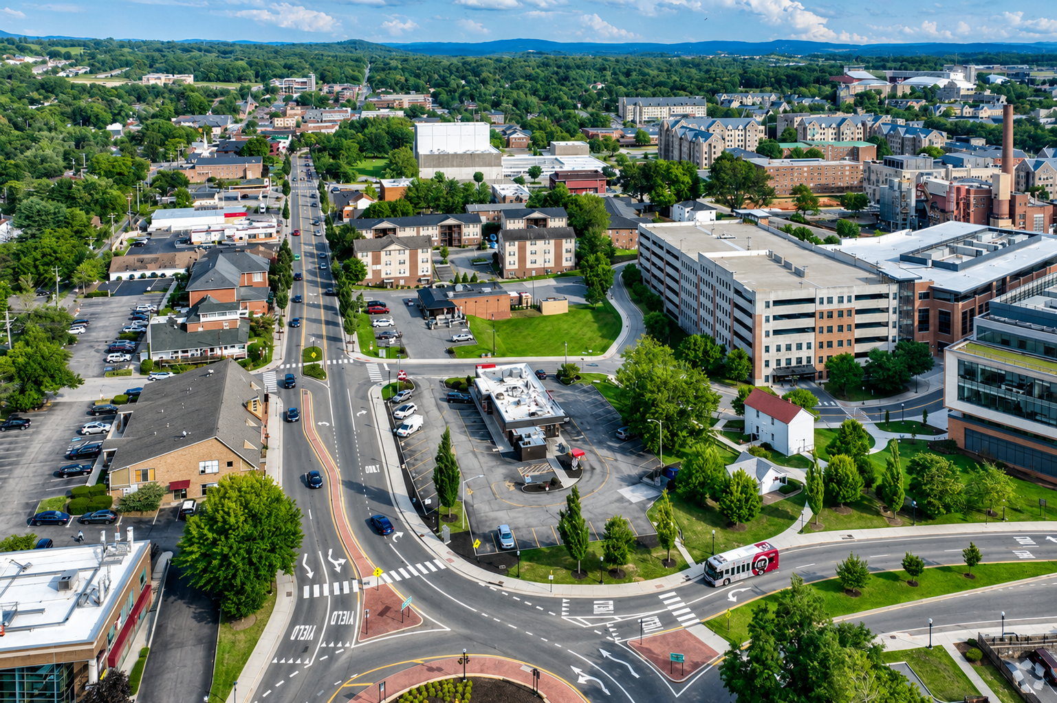 Aerial view of a city intersection with cars, buildings, and tree-lined streets under a clear sky