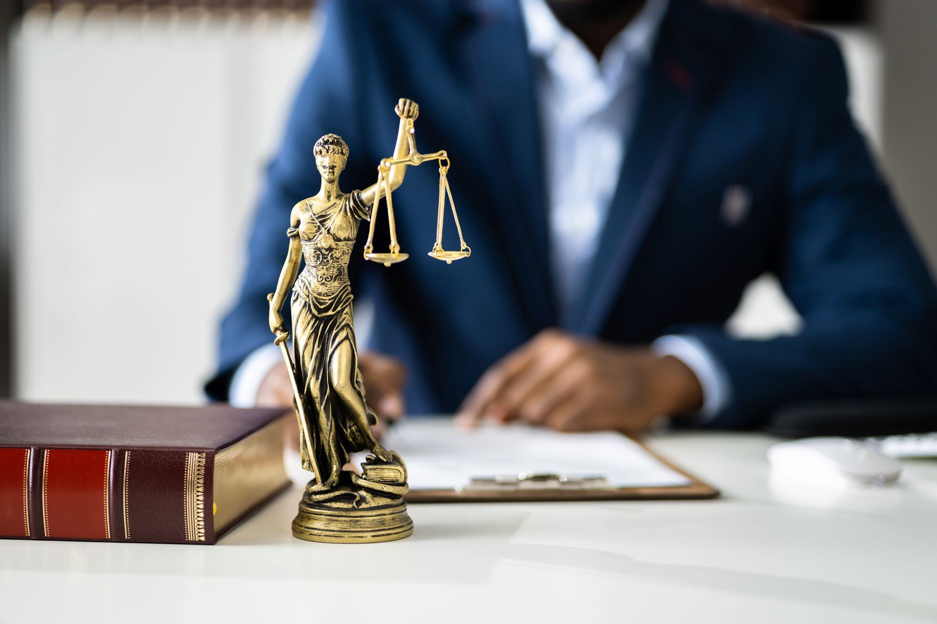 Golden Lady Justice statue on a desk beside legal books, with a blurred suited lawyer in the background