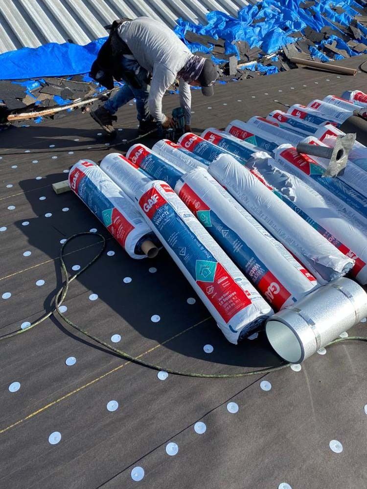 A worker installs roofing material on a flat commercial roof with multiple rolls of GAF underlayment nearby.
