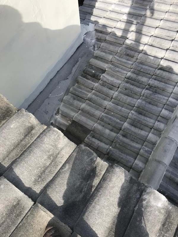 A close-up of a gray tile roof transitioning to a light-colored wall, showing roof flashing at the intersection.