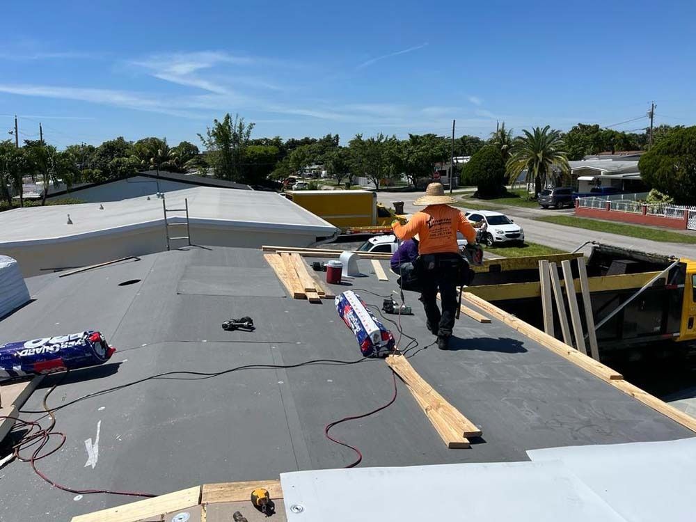 A worker in an orange shirt repairs a flat roof on a sunny day with construction materials scattered nearby.