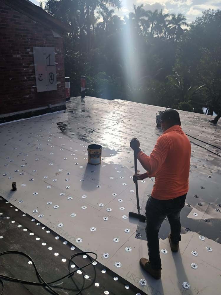 A person in an orange long-sleeved shirt and work boots uses a long-handled brush on a rooftop covered in insulation panels.