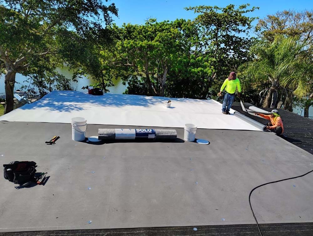 Two construction workers install a white roofing membrane on a flat, dark-colored roof surrounded by trees.