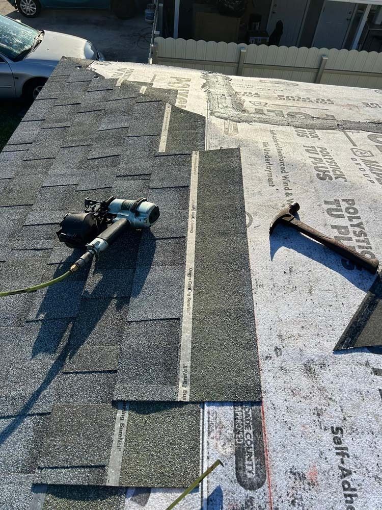 A close-up view of a roof under construction, featuring gray shingles, a nail gun, and a hammer on the underlayment.