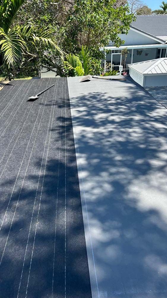A roof under repair, showing a section covered with black roofing underlayment next to a section of gray flat roofing.