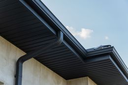 Dark metal roof edge with matching gutter and downspout against a beige exterior wall and a light blue sky.