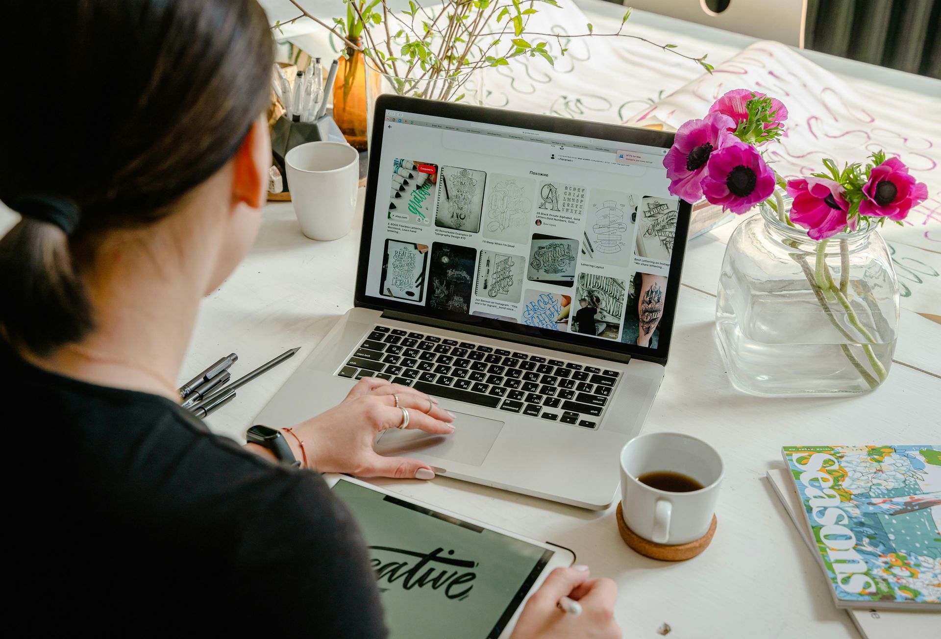 Woman working on laptop and tablet, with flowers and coffee on a white desk.