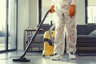 Person in protective suit vacuums a room with a yellow vacuum cleaner.